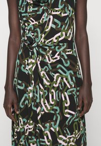 Detailní záběr na osobu s tmavou pletí, která má na sobě černé šaty s vzorem řetězu v růžové, zelené a teal barvě a uzlem v pase.