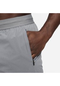 Pantalones cortos atléticos grises con una cinturilla sin costuras, bolsillo con cremallera y tejido texturizado. Presenta un ajuste relajado y un diseño transpirable.