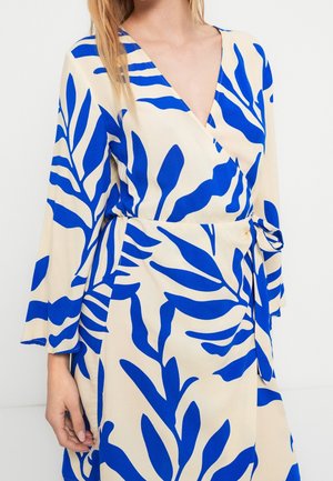 Femme portant une robe beige avec de grands motifs de feuilles bleu vif, un col en V, des manches longues et une ceinture nouée à la taille.