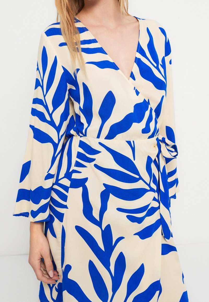 Femme portant une robe beige avec de grands motifs de feuilles bleu vif, un col en V, des manches longues et une ceinture nouée à la taille.