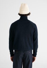 Granatowy sweter z wysokim kołnierzem i prążkowanymi mankietami, zestawiony z spodniami w paski, oraz kremową dzianinową czapką. Gładka faktura, swobodny styl.