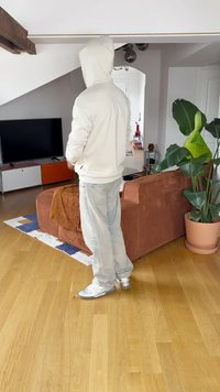 Mugav riietus, kus on heleda värvusega kapuutsiga pusa, avarad teksad ja hõbedased jalanõud, tuppa, kus on pruuni diivan ja roheline taim.