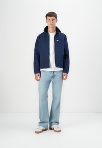 Chaqueta bomber azul marino con cierre frontal, gran cuello y dos bolsillos. Usada sobre una camisa blanca, combinada con jeans azules claros y zapatos blancos.