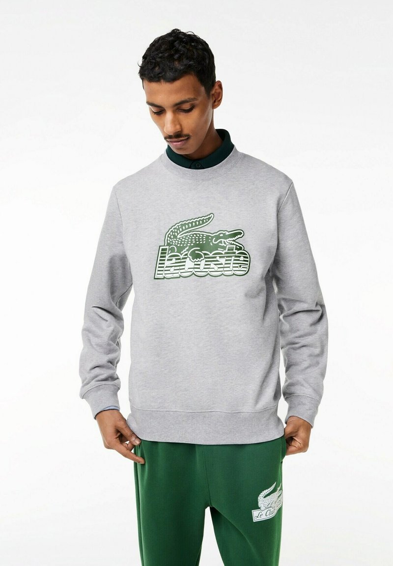 Felpa grigia con logo Lacoste verde, caratterizzata da polsini e orlo a costine. Indossata con pantaloni verdi, che mostrano uno stile casual atletico.