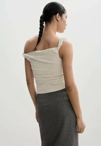 Una mujer con una coleta trenzada lleva una blusa de tirantes retorcidos color blanco roto y una falda a rayas gris oscuro, de espaldas contra un fondo sencillo.