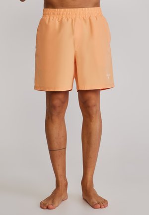 Osoba z gołymi nogami, ubrana w szorty z elastycznym pasem w kolorze brzoskwiniowym, stojąca na jednolitym, jasnym tle.