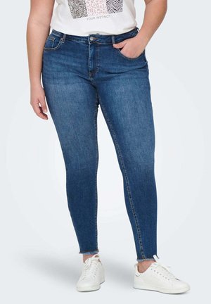 Skinny jeans in a dark blue denim. Features a fitted design, front pockets, and a raw hem. Paired with white sneakers.