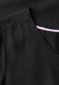 Tessuto nero con una texture morbida, caratterizzato da una scollatura a V e un evidente dettaglio in nastro di raso rosa al colletto. Cuciture pulite visibili.
