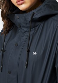 Marinblå vattentät jacka med hög krage, justerbar huva och svarta dragsnören. Har svarta tryckknappar och en diskret logga på bröstet.