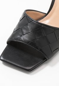 Chanclas de cuero negro con parte superior trenzada, punta cuadrada y suela de textura suave. Presenta líneas limpias y un diseño minimalista.
