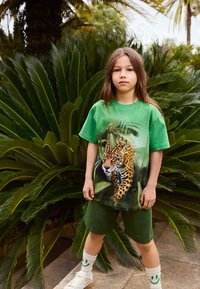 Zelena pamučna majica s dizajnom leoparda na prednjem dijelu, u kombinaciji sa zelenim kratkim hlačama i bijelim tenisicama uz čarape s osmijehom.