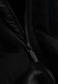 Giacca nera imbottita con zip e chiusura in velcro a contrasto, che mostra una texture liscia e sinuosa con dettagli di design minimalisti.