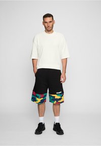 Biele oversized tričko, čierne šortky s farebným maskáčovým vzorom a logom, čierne tenisky, biele ponožky, stojí pred jednofarebným pozadím.