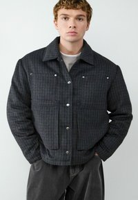 Donker geruite jas met knoopsluitingen, voorzien van twee voorkruiden, een kraag en een zachte textuur; gedragen over een lichtgekleurde shirt.