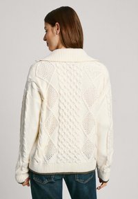 Kremo spalvos megztinis su pynėmis, su juostiniu apykakle ir tekstūruotais raštais, laisvo silueto.