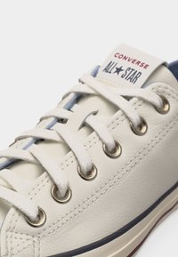 Κοντινή λήψη ενός άσπρου δερμάτινου αθλητικού παπουτσιού Converse All Star με λευκά κορδόνια, ασημένιες τρύπες και εμφανές λογότυπο της μάρκας στη γλώσσα.