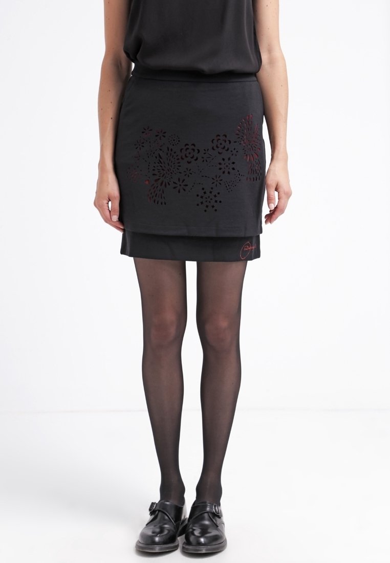 Schwarzer Rock mit lasergeschnittenen Blumenmustern entlang des Saums, über einem kürzeren Kleidungsstück getragen. Kombiniert mit schwarzen Strumpfhosen und Schuhen.