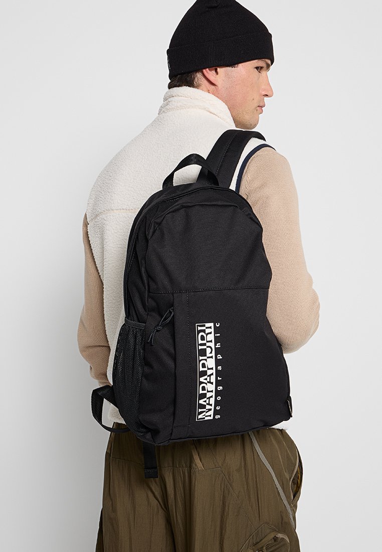 Mężczyzna w czarnej torbie napapijri geographic, beżowym swetrze, białej kamizelce polarowej, czapce beanie i oliwkowych spodniach cargo, zwrócony bokiem.