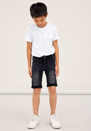 T-shirt blanche à manches courtes avec une poche poitrine, associé à un short en denim foncé avec des ourlets retroussés, porté avec des baskets blanches.
