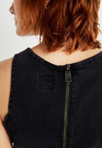 Musta denimvaate, jossa on pyöreä kaula-aukko, näkyvä kultainen vetoketju takana ja neliömäinen kangaspala.