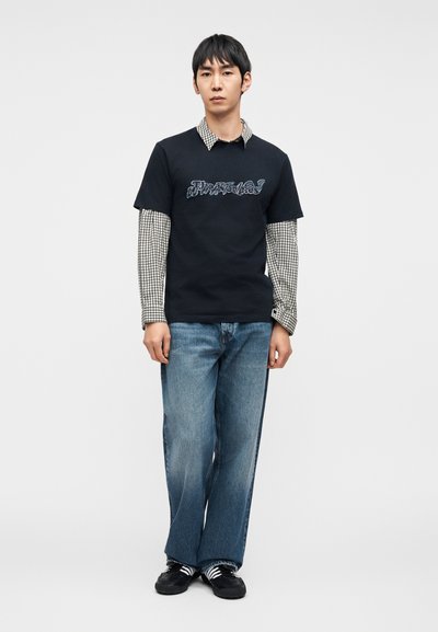 Μαύρο μπλουζάκι με μπλε γραφικό κείμενο πάνω από πουκάμισο με καρώ και μακριά μανίκια. Χαλαρό, ξεθωριασμένο μπλε τζιν και μαύρα παπούτσια ολοκληρώνουν την εμφάνιση.