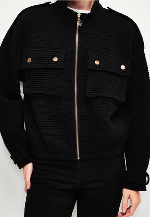 Veste noire texturée avec fermeture éclair dorée et deux poches avant à rabat avec boutons dorés, portée avec un pantalon noir.