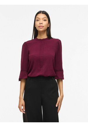 Femme portant un chemisier texturé bordeaux à manches trois-quarts et un pantalon noir taille haute, debout devant un fond blanc.