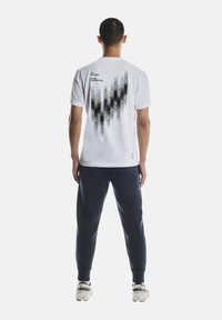 Weißes T-Shirt mit kurzen Ärmeln und schwarzem grafischem Muster auf der Rückseite, mit Text. Kombiniert mit marineblauer Hose und weißen Sportschuhen.