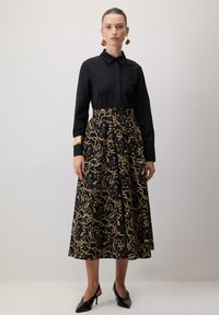 Črna dolga majica z ovratnikom v obliki konice, v kombinaciji s krilom s črno-zlatim cvetličnim vzorcem; dopolnjena z zlatim zapestnico in črnimi petkami.