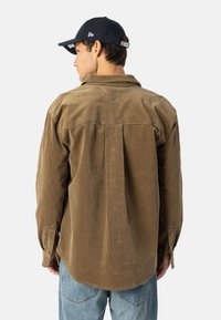 Mann iført en brun kordfløyelskjorte med bakfold, lyseblå jeans og en svart baseball-cap som vender bort fra kameraet.