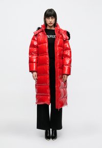 Piros, fényes felületű steppelt steppelt kabát, szőrmebéléssel ellátott kapucnival. Fekete felső és fekete bő szárú nadrág felett viselve. Fekete bokacsizma.