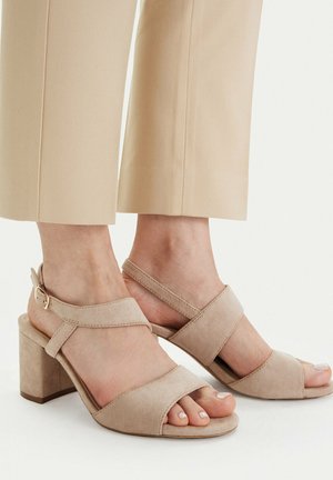Feet wearing beige suede block-heel sandals with ankle straps, paired with beige cropped trousers against a plain light background.