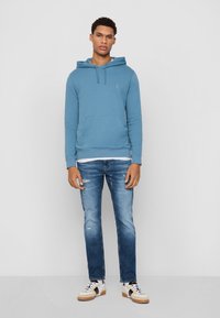 Blauwe katoenen hoodie met een voorzak, gecombineerd met lichtblauwe versleten denim jeans en witte sneakers met zwarte accenten.