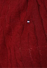 Maglione rosso lavorato a maglia con motivo a treccia e un piccolo logo blu, bianco e rosso. La texture appare spessa e calda.