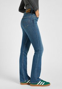 Blå denimjeans med slim fit och lätt flare i botten. Har en brun läderlinning med "Lee"-logotyp. Matchas med gröna sneakers.