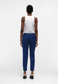 Vrouw met kort krullend haar, gekleed in een witte mouwloze top, blauwe slim-fit broek en zwarte hoge hakken, staand met haar rug naar de camera op een witte achtergrond.