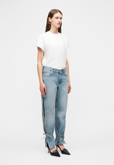 Γυναίκα που φοράει λευκή κοντομάνικη μπλούζα, ανοιχτό μπλε τζιν με πλαϊνές σχισμές και δεσίματα στους αστραγάλους, και μαύρα ψηλοτάκουνα με μυτερή μύτη, στέκεται μπροστά σε απλό φόντο.