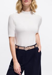 Mujer vestida con una blusa de punto blanca de manga corta metida en pantalones oscuros a rayas con un cinturón beige y hebilla plateada, de pie con una mano en el bolsillo.