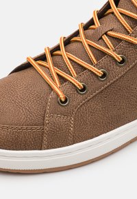 Zapato marrón texturizado con cordones a rayas naranjas y negras, ojales metálicos y una suela de goma blanca, que presenta un sutil detalle de costura.