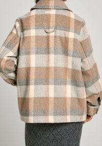 Persona vestida con una chaqueta de cuadros en tonos beige, gris y crema, y una falda oscura texturizada, vista desde atrás sobre un fondo liso.