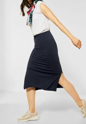 Vrouw met witte top, donkere knielange rok met zijsplit, beige sneakers en een kleurrijke sjaal lopend tegen een effen achtergrond.
