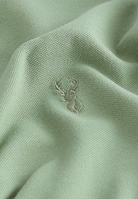 Svijetlo zeleni pleteni materijal s finom teksturom, s izvezenim dizajnom jelena u malo tamnijoj nijansi. Tkanje je gusto i ujednačeno.
