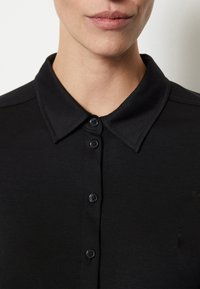 Camisa negra de botones con cuello, cinco botones negros, textura suave y un diseño ajustado, mostrando el escote y la parte superior del pecho.