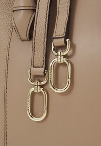 Beige nahka laukku, jossa on kaksi kultavaa metalliklipsiä, jotka on kiinnitetty teksturoituihin nahkahihnoihin, joissa on tumman kontrastivärinen ommel.