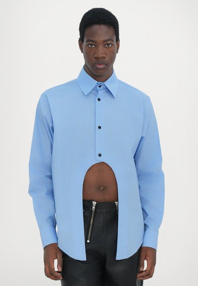 Chemise bleu clair à boutons avec une grande découpe au niveau du torse, manches longues et boutons noirs, portée avec un pantalon zippé noir.