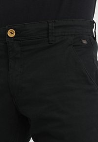 Pantalones negros con un botón beige, bolsillos delanteros y presillas para cinturón, llevados con una blusa negra metida por dentro.