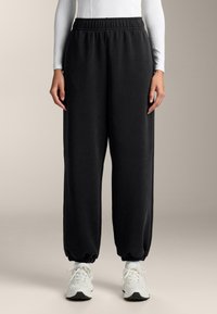 Čierne tepláky vyrobené z mäkkej textílie, s elastickým pásom a manžetami, uvoľneného strihu a bočnými vreckami.