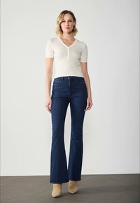 White, fitted short-sleeve top with a v-neck and button accent, paired with dark blue flared jeans and light beige ankle boots.
