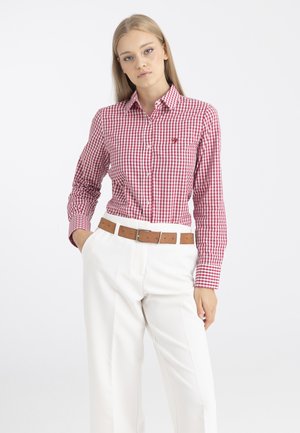 Femme blonde portant une chemise à manches longues à carreaux rouges et blancs rentrée dans un pantalon blanc avec une ceinture marron, debout avec une main dans la poche.