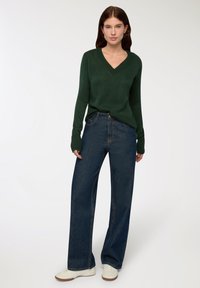 Ciemnozielony, prążkowany sweter z dekoltem w serek zestawiony z wysokim stanem, szerokimi spodniami dżinsowymi w kolorze niebieskim. Model nosi białe sneakersy. Prosta, codzienna stylizacja.